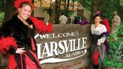 Two women in saloon attire smile while standing next to a sign that reads 'Welcome to Liarsville, Alaska"
