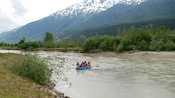 A small group paddling down a river in an inflatable raft alongside a snowcapped mountain