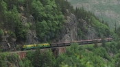 A vintage train travels alongside a verdant mountain face