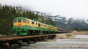 A vintage train rolls along the tracks over a lake through mountainous terrain