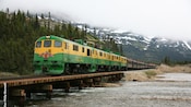 A vintage train travels between a river and a mountainside