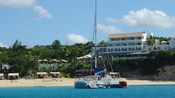 Un catamarán anclado cerca de una costa de una playa tropical con cabañas, un restaurante y un hotel