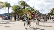 A girl on a bike smiles at the camera as other bicyclists ride behind her, next to palm trees and a beach