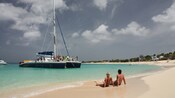 A couple lounges on the sand of a tropical beach while a catamaran is anchored nearby