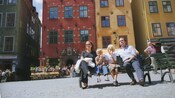A family sits on a bench, eating ice cream, in front of a café and brightly colored old buildings