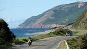 A car and a motorcycle driving along a winding street down a hill alongside a large body of water