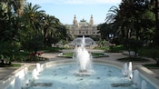 A large, ornate building overlooks a large circular fountain, several planters and 2 fountains