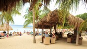 A sandy beach filled with chairs, people, palm trees and tropical thatch umbrellas for sun protection