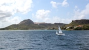 A lone sailboat sits on the ocean, not far from the islands lush, mountainous shoreline