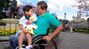 A young boy with a plush toy sits on his father's lap, who is using a wheelchair at Disneyland Park
