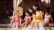 A mom and 3 young girls sit at the edge of the pool and splash their feet
