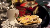 A plate with Christmas cookies next to a mug of chocolate topped whip cream near presents