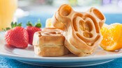 A Mickey Mouse shaped waffle propped up by another waffle on a plate with strawberries and an orange wedge