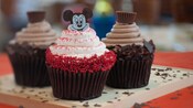 3 chocolate cupcakes, 1 with white frosting and red sprinkles topped with a Mickey Mouse image