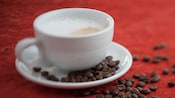 Cup of cappuccino with coffee beans scattered in saucer and on table