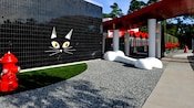 Un pared exterior negra brillante adornada con la cara de un gato al lado de un área de grava con un hueso gigante de perro y un área de césped con una boca contra incendios