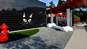 Uma parede externa em preto brilhante decorada com uma cara de gato grande, ao lado de uma área com cascalho com um osso de cachorro gigante e outra área coberta de grama com um hidrante.