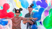 A father and son wearing Mickey Mouse ear hats laugh as they’re surrounded by Mickey Mouse balloons