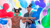 A father and son wearing Mickey Mouse ear hats laugh as they’re surrounded by Mickey Mouse balloons