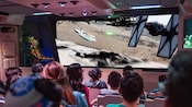 A seated audience watches imagery on a screen depicting an aerial chase in which a TIE fighter pursues and fires blasters at the Millennium Falcon
