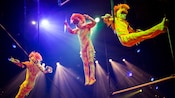 Three acrobats in orange monkey outfits perform on gymnastic rings