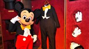 Mickey Mouse frente a un armario lleno de ropa, que incluye su esmoquin y sus guantes blancos