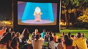 Visitantes sentados na grama assistindo ao filme A Bela e a Fera, à noite