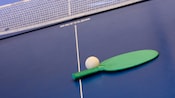 Close-up of a green ping-pong paddle and a white ball on a blue table