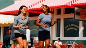2 mujeres disfrutan de correr mientras pasan frente a una tienda con un toldo a rayas rojas y blancas