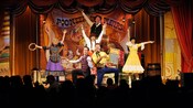 A group of performers in pioneer clothing pose on a stage with a sign that reads Pioneer Hall Players