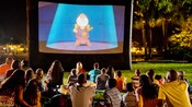 Um grupo de visitantes reunido em um jardim para assistir à projeção de um filme da Disney