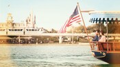 A couple glides past Magic Kingdom park aboard an American flag bearing ferryboat