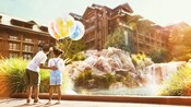 A young girl holds 3 Disney themed balloons as her older brother taps her back and points to the Resort’s accommodations