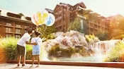 Uma menina segura 3 balões temáticos da Disney enquanto o irmão mais velho toca em suas costas e aponta para as acomodações do Resort