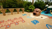 Vegetação em volta de um playground com inspiração maia, brinquedos com tema de floresta e uma caixa de areia com uma grande estátua oca de uma cabeça de maia