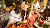 Una mujer y una niña sostienen binoculares sentadas en la mesa de un restaurante con platos de comida