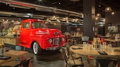 Restaurante bajo techo muestra un camión vintage con canastas de comida, mesas con bebidas, utensilios y sillas