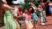 A small girl wearing a lei and grass skirt smiles as hula dancers teach her family the hula dance