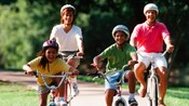 Una familia de 4 pasean en sus bicicletas con cascos por un sendero bordeado de árboles