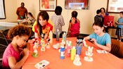 Un miembro del elenco observa a 2 niños pintando figuras de Disney en una mesa de manualidades