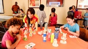 Un Membre de l’équipe surveillant 2 enfants en train de peindre des figurines Disney sur une table d’art plastique