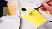 A brush held by a Guest painting a cardboard birdhouse yellow