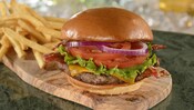 Un cheeseburger garni d’une boulette de viande, de bacon, de laitue, de tranches de tomate, d’oignons violets et de fromage cheddar près d’une pile de frites