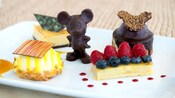 A pastry topped with blueberries and raspberries near a chocolate topped pastry, a rounded pastry topped by a chocolate, a triangular pastry and a chocolate figurine of Mickey Mouse
