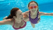 A mother swims underwater with her daughter who’s wearing goggles