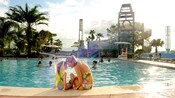 A little girl wearing a swimsuit with large fairy wings sitting with her mother at the edge of a swimming pool at a Disney Resort hotel