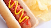 A plump hot dog with mustard and ketchup next to a side of potato chips