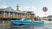 An amphibious car drives through the water with a family in the backseat with a pier behind it