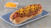 A hot dog covered in chili and shredded cheddar cheese on a plate near a dish of cheddar cheese