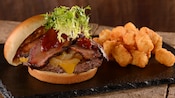 Una hamburguesa de carne cubierta con queso, cebollas, rodajas de falda de res y lechuga, al lado de una pila de croquetas de papa